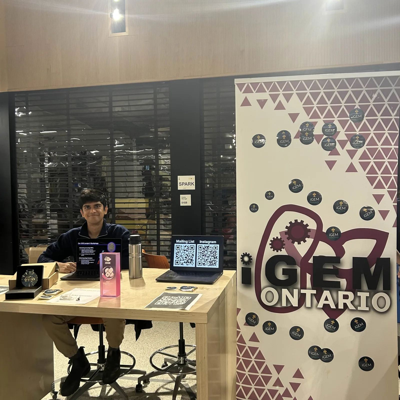Team member staffs the iGEM Toronto table beside the 2025 project display and awards, January 2026